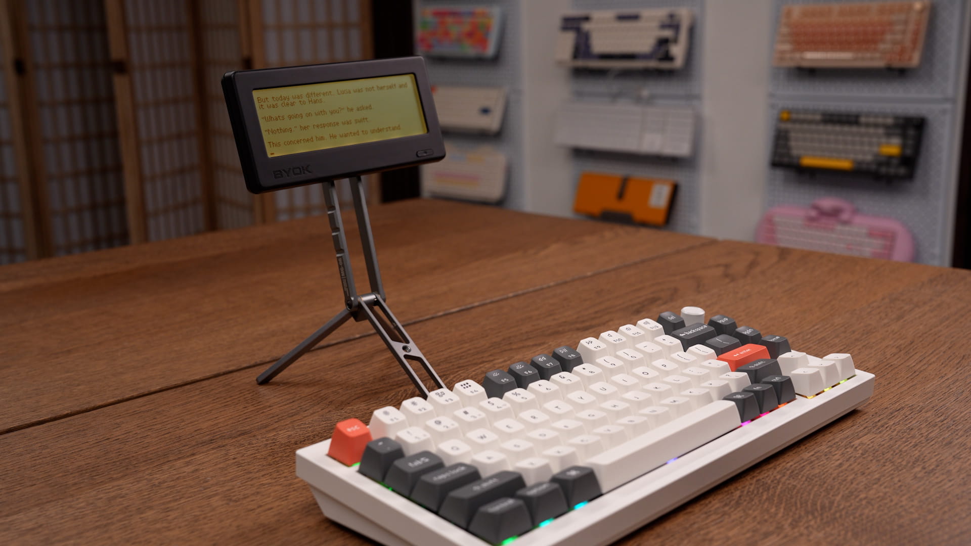 BYOK writing device on a metal tripod stand behind a Keychron Q1 Ultra keyboard, with a wall of keyboards in the background